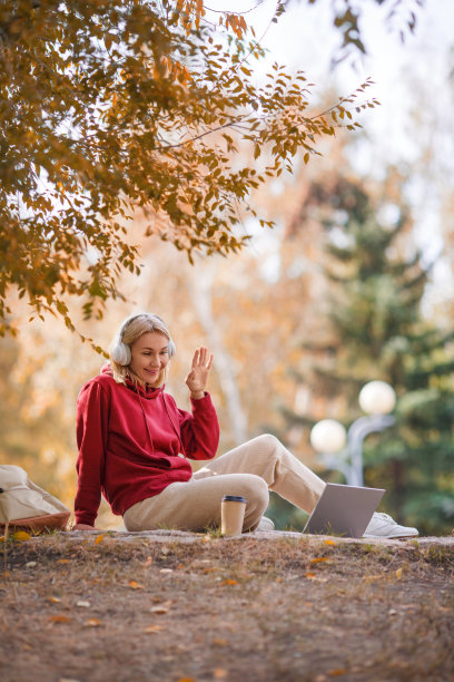 视频电话。戴着耳机的微笑女子坐在笔记本电脑前，端着咖啡，在秋天的户外公园里与人打招呼，垂直照片，选定焦点。图片下载
