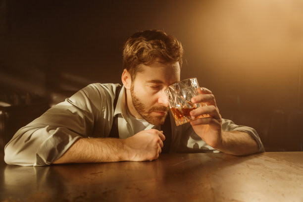 一个男人拿着一杯威士忌的大气照片。一个男人喝得酩酊大醉，坐在桌旁，他把酒杯放在前额上。那人闭上了眼睛，他在酗酒中昏倒。图片下载