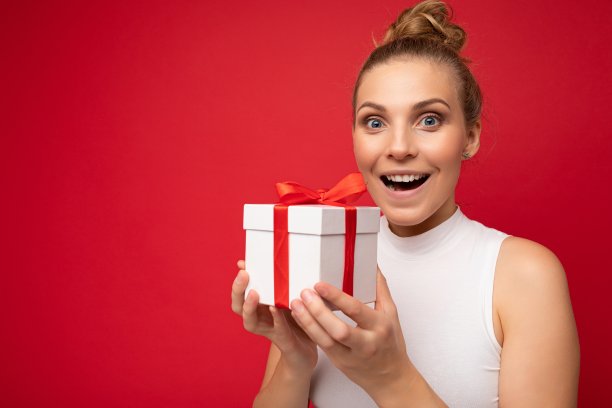 照片拍摄的相当积极的惊讶年轻的金发女人孤立在彩色背景墙穿着时髦的服装看拿着礼品盒，看着相机图片下载