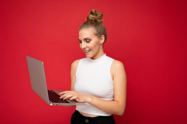 侧面照片照片美丽的微笑快乐的金发年轻女子与聚集头发穿白色t恤使用电脑笔记本电脑打字键盘上看着上网本显示器屏幕隔离在红色的墙背景图片下载
