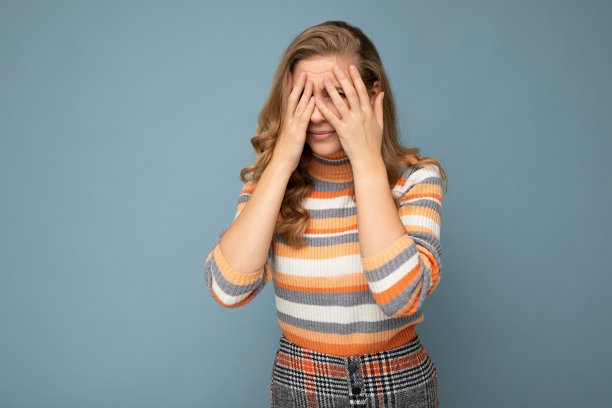 年轻的金发美丽的卷发女人的肖像，真诚的情绪穿着条纹毛衣孤立在蓝色的背景，空白的空间和遮住眼睛图片下载