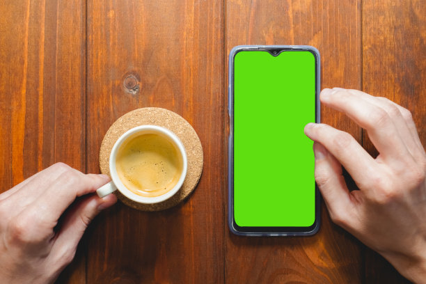 模型形象的手拿着手机与空白的绿色屏幕与咖啡杯在木桌上。俯视图图片下载