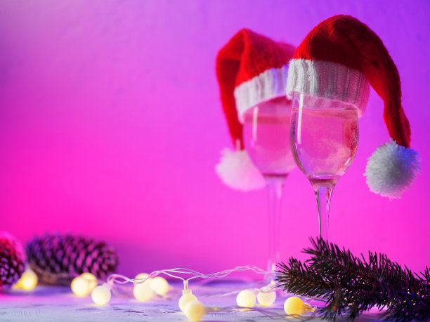 两杯在圣诞老人的圣诞帽里的起泡葡萄酒或香槟。霓虹时尚的紫色与粉红色的copyspace背景。圣诞锥和圆形花环图片下载