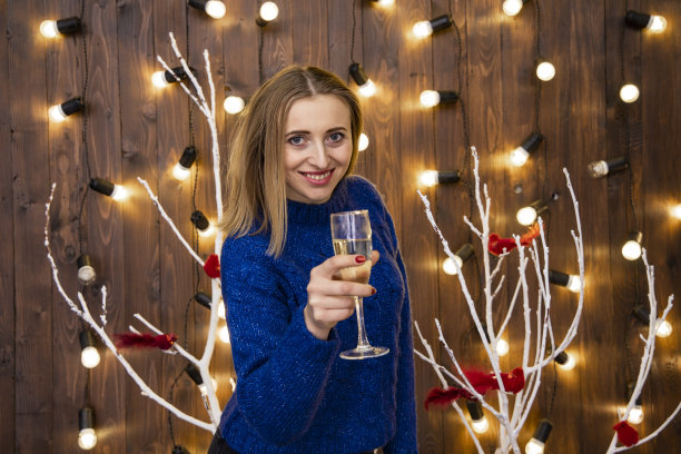 快乐女人笑脸女性肖像圣诞夜在工作室室内用一杯香槟和花环灯黄色照明背景图片下载