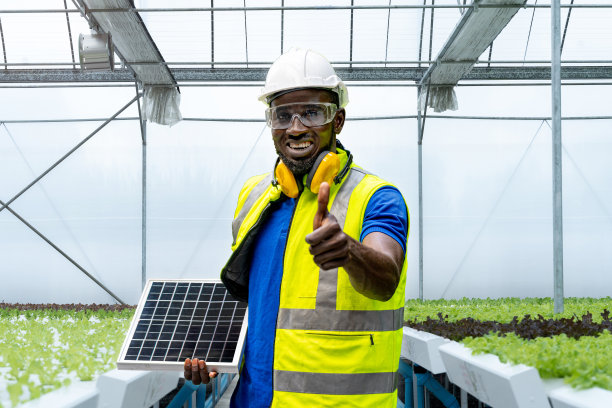 工人穿着工作服、戴着安全帽站在温室前，手持太阳能电池板。工人为农业植物和蔬菜建立了一个水培系统。图片下载