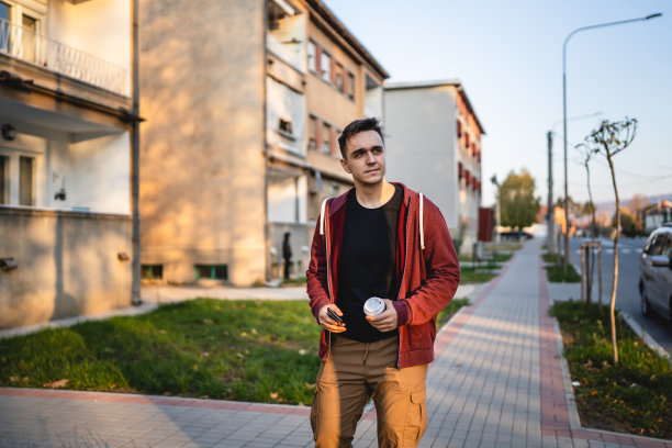 前面的看法，一个成年年轻的白人男子走在大厦前的城镇在秋天的一天，拿着手机和一杯咖啡的半长真人复制空间看向一边图片下载