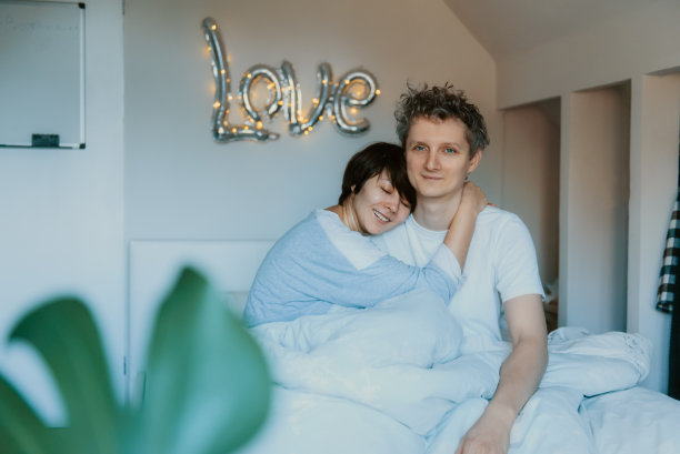 一对情侣穿着睡衣在卧室的床上玩耍，墙上的背景上写着“爱”。他们拥抱着，笑着。幸福的家庭早晨在家里。情人节。有选择性的重点。图片下载