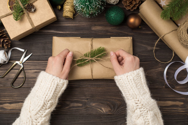 第一个人俯视图照片的女性手在毛衣绑线蝴蝶结在工艺纸礼品盒装饰与松枝绿色金球和手工工具在孤立的木制书桌背景图片下载