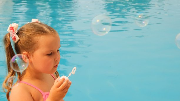 快乐的4-5岁小女孩，扎着马尾辫，穿着粉红色的泳衣，在暑假的游泳池里吹肥皂泡球图片下载