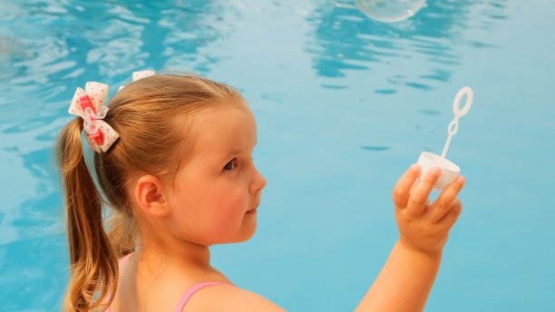 可爱的4-5岁小女孩，扎着马尾辫，穿着粉红色的泳衣，在暑假的游泳池里吹肥皂泡图片下载