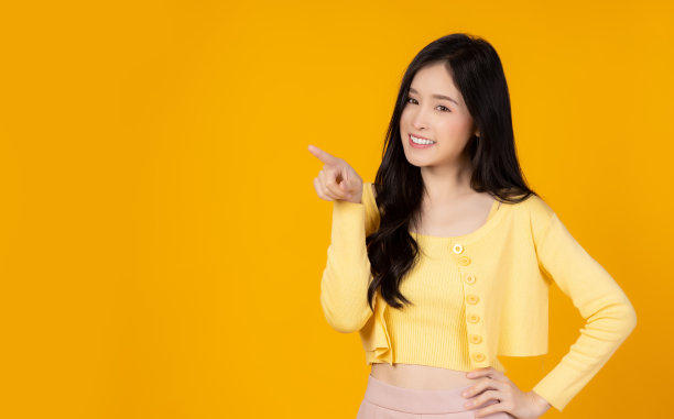 快乐的年轻亚洲女人指向复制空间，微笑的脸和幸福的看着相机漂亮的女孩表现出满意的产品，用于广告与黄色的背景，复制空间图片下载