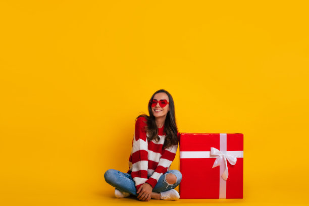 完整的照片，一个兴奋的美丽时尚的黑发女人戴着太阳镜，她坐着，摆着一个红色的大礼盒图片下载