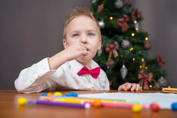 可爱严肃的男孩，穿着白色衬衫，打着粉色领结，在平安夜在家给圣诞老人写信，想着礼物，吃着甜甜的糖果图片下载