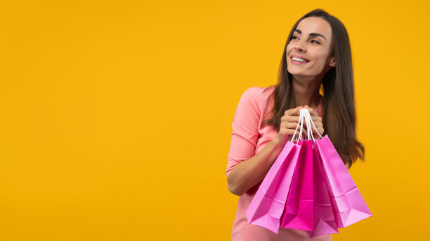 迷人自信的年轻女子肖像在时尚的淡粉色连衣裙摆在明亮的黄色背景下，手里拿着一堆购物袋。图片下载