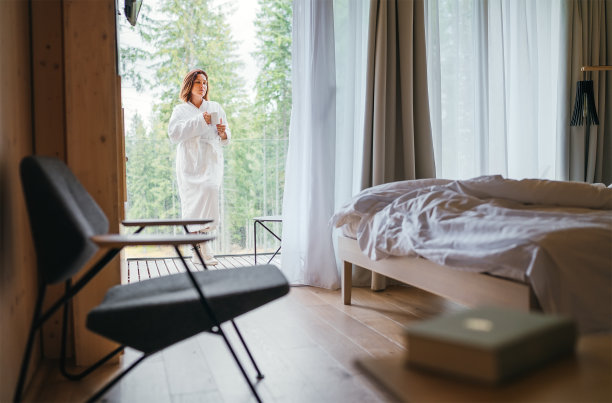 穿着白色浴衣的女人站在森林房子的阳台上，享受着新鲜的空气和早晨的茶杯。在一个斯堪的纳维亚的室内设计室视图与一个没有整理的床。舒适的生活理念形象。图片下载