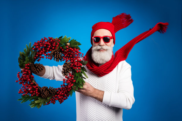 照片相当迷人的成熟男子穿着白色套衫红色头饰持圣诞花环孤立的蓝色背景图片下载