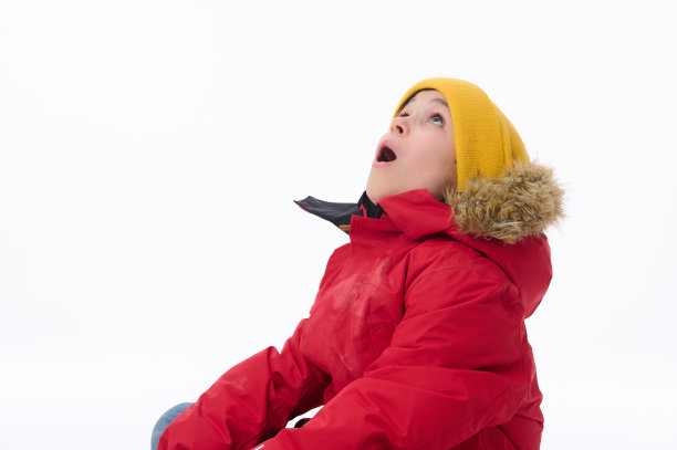 一个英俊的、惊讶的、惊讶的欧洲小学年龄的男孩在明亮的黄色羊毛帽子和红色的皮大衣抬头看着张开嘴对着白色的墙壁背景与拷贝广告空间图片下载