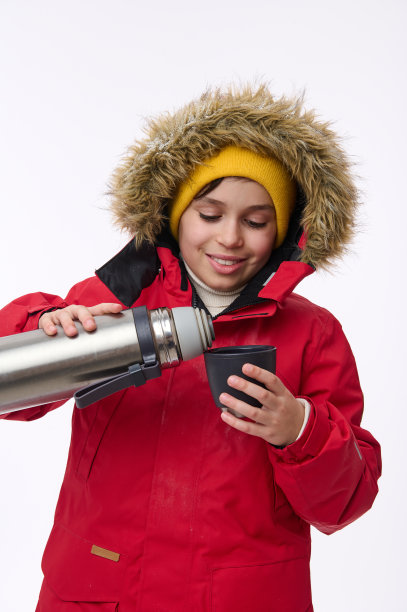 垂直肖像一个可爱的学龄白人男孩冬季旅行者冒险家倒热饮从保温瓶到一个杯子，孤立的白色背景与广告的复制空间图片下载