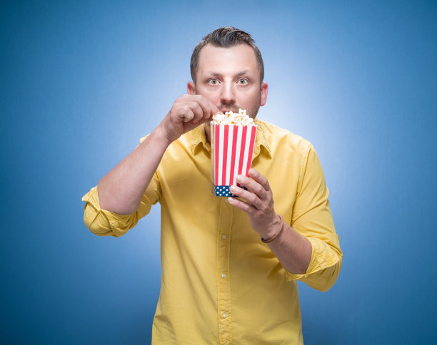 拿着爆米花，穿着黄色衬衫，在蓝色背景下看恐怖电影。害怕的年轻人。周末时段-观影概念图片下载