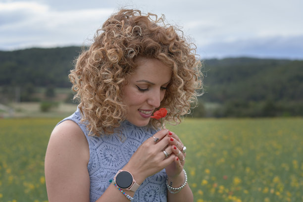 一个年轻的女孩，她有一头金色的卷发，手指上戴着一朵红花，正在闻花。图片下载