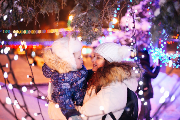 一名年轻女子抱着小女孩在圣诞树下，在圣诞市场装饰的节日广场上图片下载