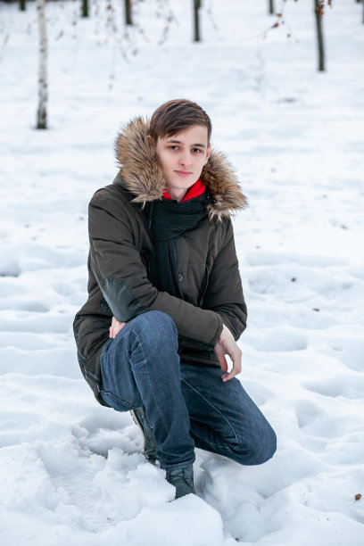 一个年轻人穿着冬季皮草夹克坐在公园里，背景是白雪覆盖的地面图片下载