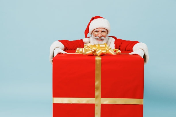 全身上下的圣诞老人50多岁，戴着圣诞帽，穿着红色西装，站在大礼物盒旁边，孤立在朴素的蓝色背景下，工作室新年快乐，2022年庆祝，圣诞快乐的节日概念图片下载