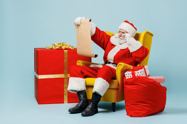 全身上下的圣诞老人50多岁，戴着圣诞帽，穿着红色的衣服，坐在扶手椅上，旁边是礼物袋，读着信，孤立在纯蓝色背景的工作室里。新年快乐，2022年圣诞快乐。图片下载