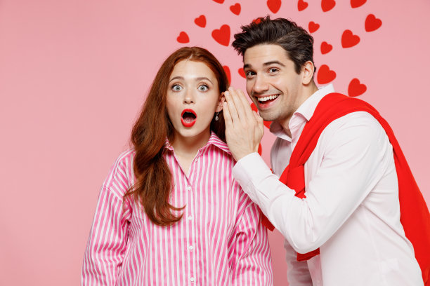 年轻震惊的夫妇，两个朋友，女人，男人，穿着休闲衬衫，在她的手背后低语八卦和秘密，孤立在素色淡粉色的背景情人节生日节日派对的概念。图片下载
