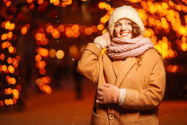 在夜晚的花环灯的背景下，年轻的女人穿着冬季风格的衣服。图片下载