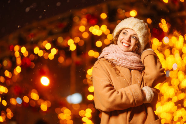 在夜晚的花环灯的背景下，年轻的女人穿着冬季风格的衣服。图片下载