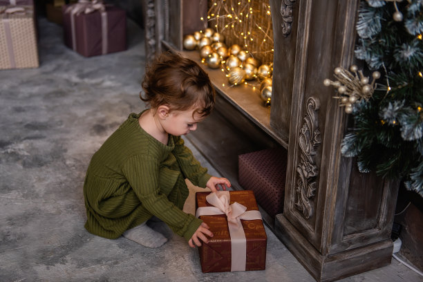 小女孩穿着温暖的绿色衣服，手捧金球，站在壁炉旁的圣诞树旁图片下载