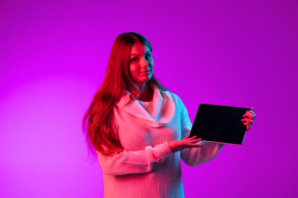 一个快乐的年轻女孩穿着暖和的针织衫，用平板电脑，在霓虹灯下紫色的工作室背景上孤立的小装置。图片下载