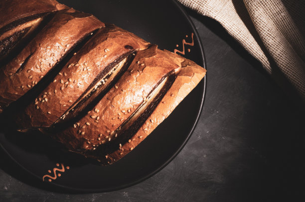 不同寻常的两色新鲜面包放在陶瓷盘子里，一张编织餐巾放在黑色背景上，照片在强光下低调拍摄，特写。图片下载