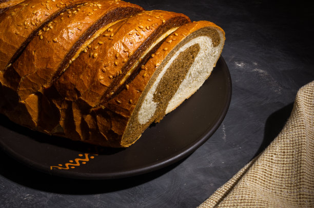 不同寻常的两色新鲜面包放在陶瓷盘子里，一张编织餐巾放在黑色背景上，照片在强光下低调拍摄，特写。图片下载