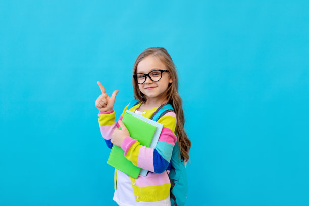 一张可爱的小女孩的画像，她戴着眼镜，穿着条纹夹克，手里拿着笔记本和课本，背着背包。教育的概念。蓝色背景，放置文本。图片下载