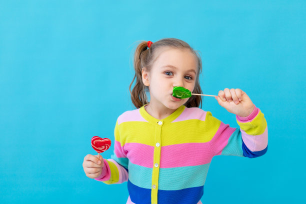 一个可爱的小女孩的肖像，穿着条纹夹克，拿着棒棒糖。那孩子吃了又舔了一根大棒棒糖。糖果和糖果的概念。照片工作室，蓝色背景，放置文本图片下载