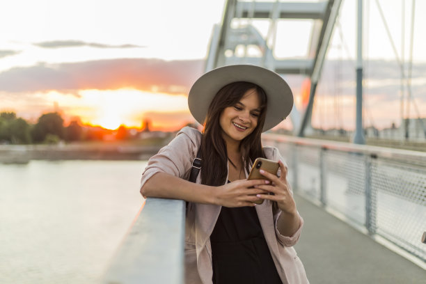 一个年轻的微笑的女人倚在桥栏上，用手机打字。一辆电动滑板车停在她旁边。河流、日落和现代桥的部分在背景中占主导地位。图片下载