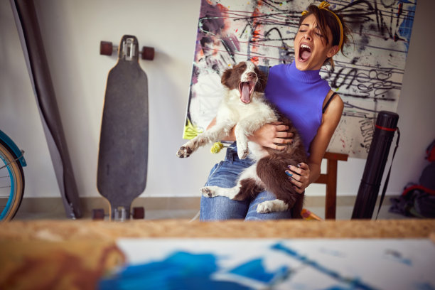 一位年轻的女艺术家和她的狗在她的工作室里玩得很开心，正在打哈欠。艺术，绘画，友谊，工作室图片下载