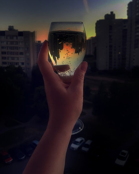 一个女人手里拿着一杯香槟的特写镜头，模糊的城市背景。在一杯透明的葡萄酒中，建筑的倒影和令人惊叹的日落。真理在酒里图片下载