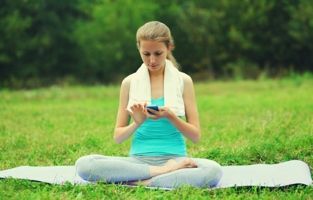 健身妇女与智能手机在垫子上的草地在夏季公园图片下载