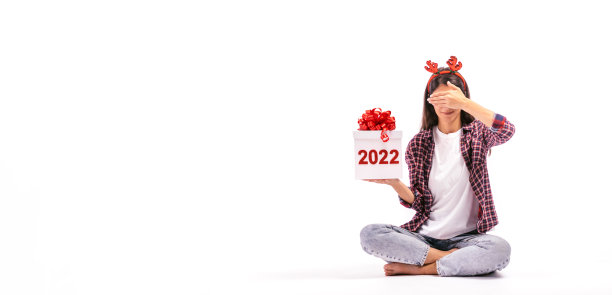 年轻的黑发女子坐在白色背景上，手里拿着一个写着“2022”的礼品盒，用手遮住眼睛。2022年新年的概念。图片下载