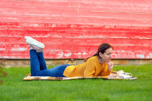 一个穿着黄色毛衣的女孩躺在草坪上，手里拿着一台笔记本电脑图片下载