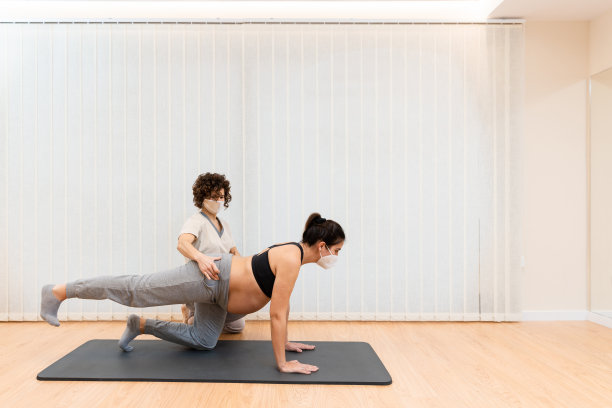 一个带着面罩的孕妇跪在垫子上做普拉提，在理疗师的帮助下有复制空间图片下载