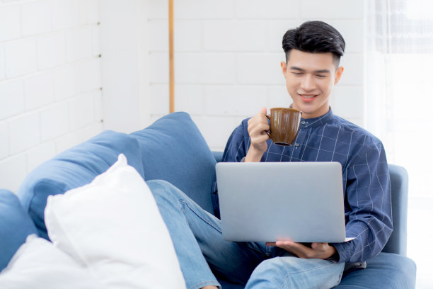 年轻的亚洲商人在客厅的沙发上用笔记本电脑在线在家工作，自由商人在沙发上用笔记本电脑喝咖啡，新常态，生活理念。图片下载