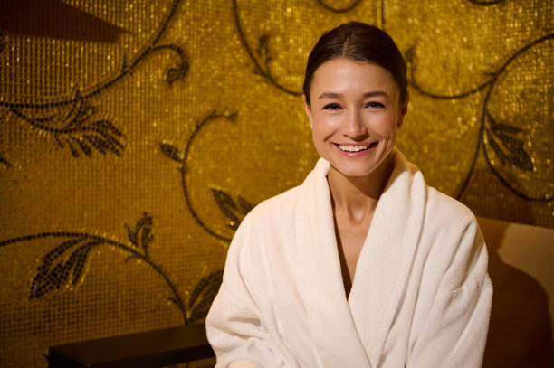 近距离拍摄的照片中，一位身着白色毛巾布浴袍的欧洲中年妇女坐在健康水疗中心休息区的扶手椅上，对着镜头露出了露齿的微笑图片下载