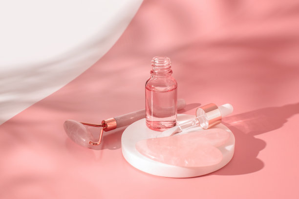 按摩石英滚轮和粉色玻璃瓶化妆品透明凝胶液体在粉红色的背景。滴管瓶，透明质酸，油脂，血清。面部按摩和放松。刮痧按摩工具。图片下载
