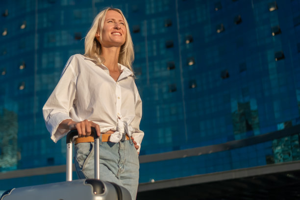 一个年轻快乐的金发女人的肖像，在白色衬衫和牛仔裤与一个手提箱在她的手的背景下，一个现代时尚的玻璃酒店在夕阳的光线与复制空间图片下载