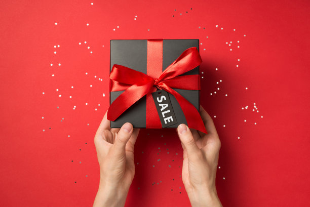 第一人称俯视图照片的手拿着黑色礼品盒与红色丝带蝴蝶结和标签闪亮的亮片在孤立的红色背景上与文本的价格标签图片下载