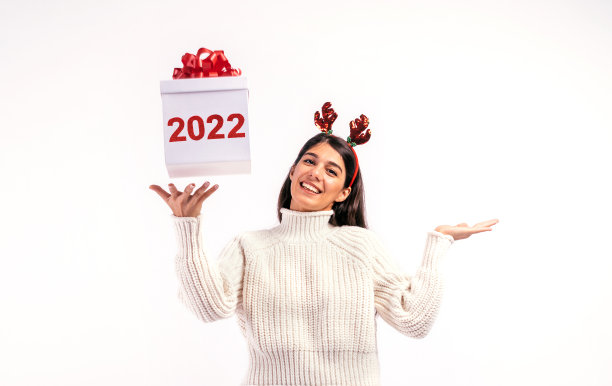 写着“2022”的白色礼盒落入女孩手中。女孩站在白色的背景上，穿着白色的毛衣，头上装饰着圣诞鹿角。2022年新年的概念图片下载